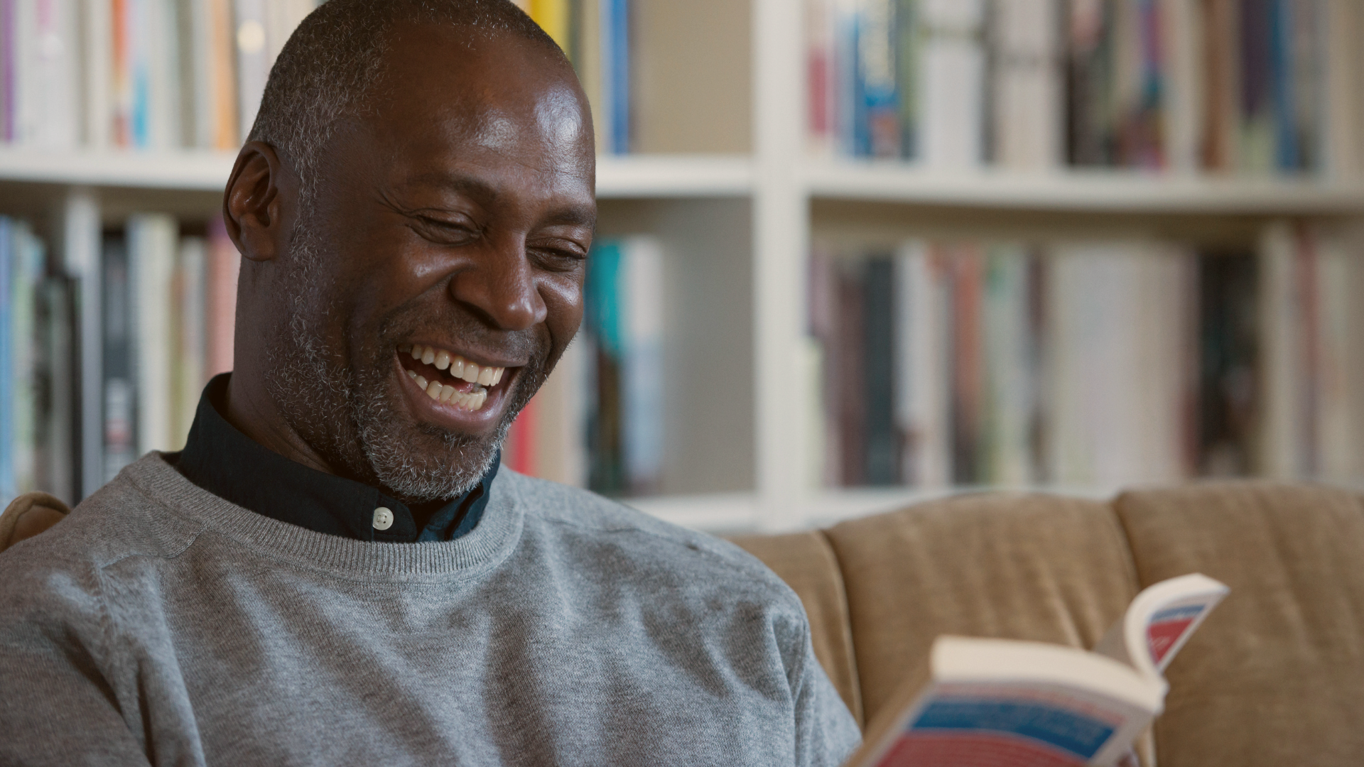 Untitled design (23) Man laughs as he's reading an entertaining, humorous book to improve his mental health.