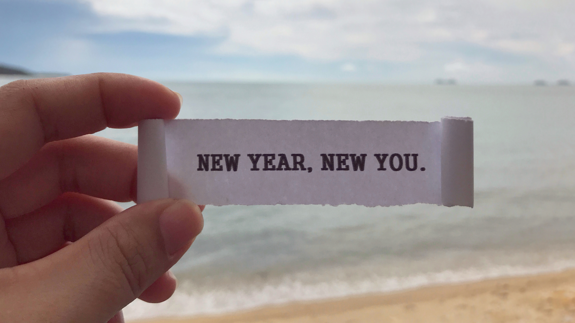 person holding a piece of paper that says new year new you with the beach in the background